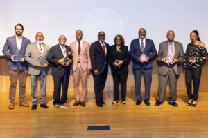 diverse group of eight event honorees holding matching glass awards, standing together on a stage in front of a projection screen displaying a "Legacy Business" logo.