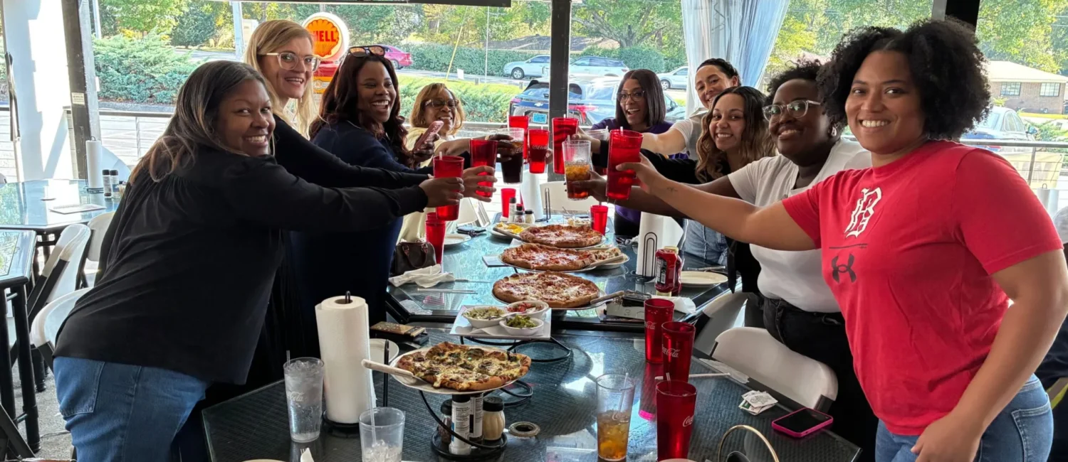 A group of diverse women smiling and raising their glasses in a celebratory toast while gathered around a long table on a restaurant patio. The glass table is covered with several pizzas, salads, and beverages. In the background, a television screen is mounted on the wall, and large windows show a view of the street, including a Shell gas station sign.