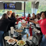A group of diverse women smiling and raising their glasses in a celebratory toast while gathered around a long table on a restaurant patio. The glass table is covered with several pizzas, salads, and beverages. In the background, a television screen is mounted on the wall, and large windows show a view of the street, including a Shell gas station sign.