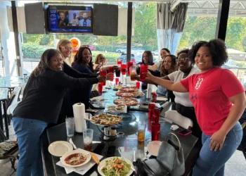 A group of diverse women smiling and raising their glasses in a celebratory toast while gathered around a long table on a restaurant patio. The glass table is covered with several pizzas, salads, and beverages. In the background, a television screen is mounted on the wall, and large windows show a view of the street, including a Shell gas station sign.