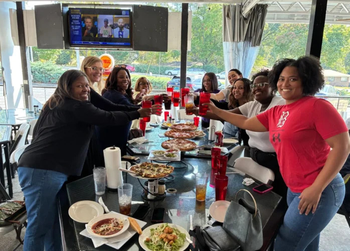 A group of diverse women smiling and raising their glasses in a celebratory toast while gathered around a long table on a restaurant patio. The glass table is covered with several pizzas, salads, and beverages. In the background, a television screen is mounted on the wall, and large windows show a view of the street, including a Shell gas station sign.